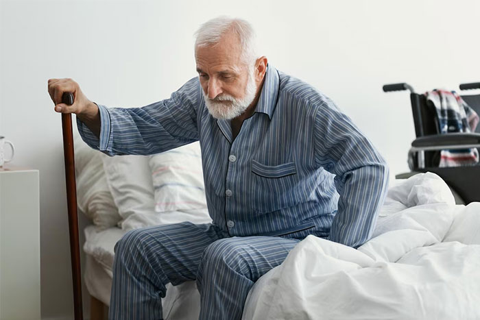 Elderly man in striped pajamas sitting on bed with cane, representing people who work in strangers’ homes seeing unforgettable things.