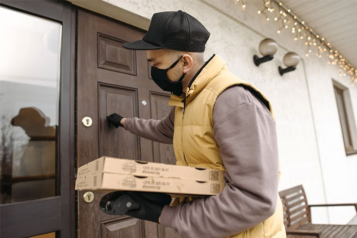 Delivery worker wearing a mask and gloves, holding pizza boxes while knocking on a stranger’s home door.