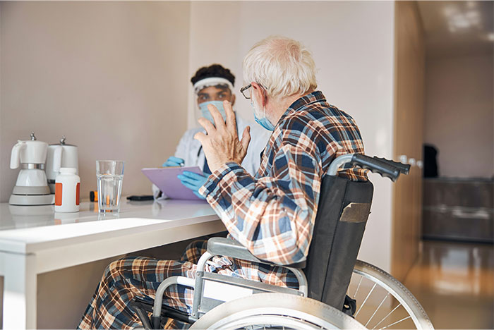 Health worker in a mask consulting an elderly man in a wheelchair inside a stranger’s home, sharing unforgettable moments