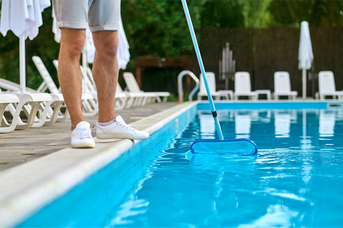 Person working in strangers’ homes cleaning a private swimming pool with a net skimmer on a sunny day.