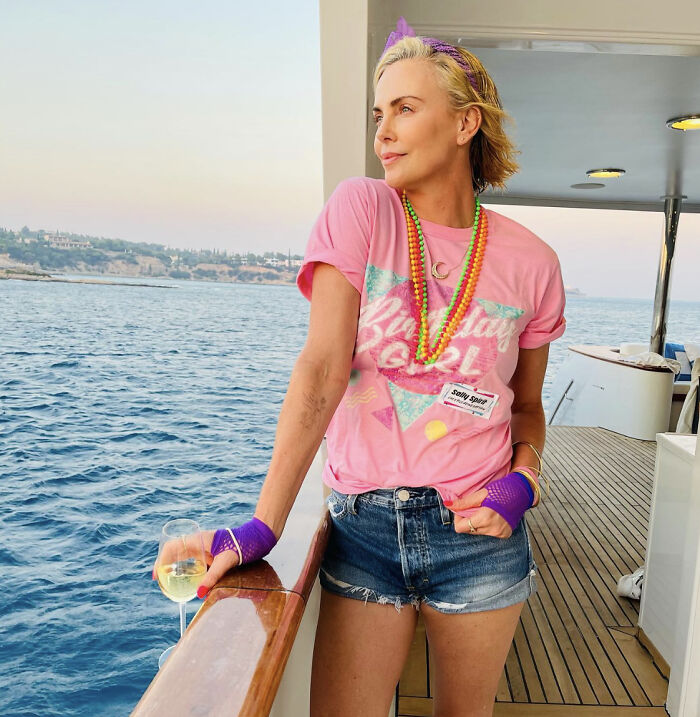 Charlize Theron wearing pink shirt and denim shorts, relaxing on a boat with a glass of wine by the sea.