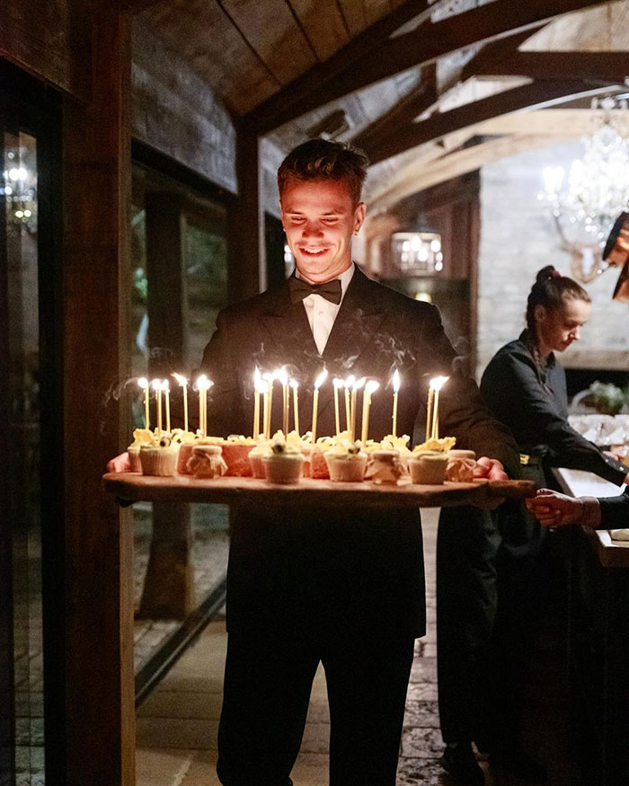 Young man in tuxedo holding birthday cupcakes with lit candles at David Beckham 50th birthday party family event.