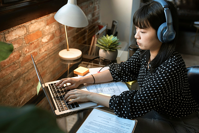 Woman wearing headphones working on laptop at office desk, depicting coworker seeking peaceful 9-to-5 experience.