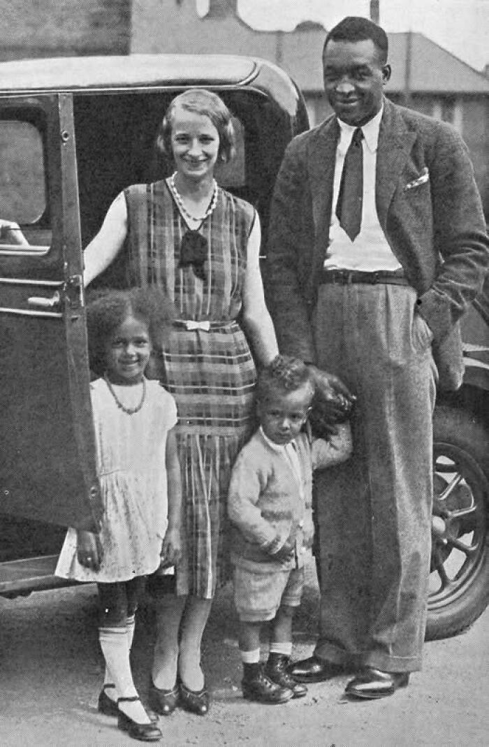 Black and white photo of an interracial family from the early 20th century standing beside a vintage car, unusual historical fact.