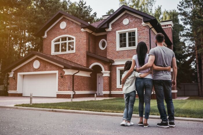 Family of three standing outside a large brick house in a suburban neighborhood symbolizing middle class living.