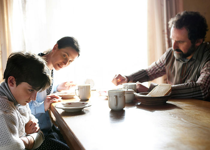 Teenager sitting apart from family at table, showing tension with stepmom after graduation invitation conflict.