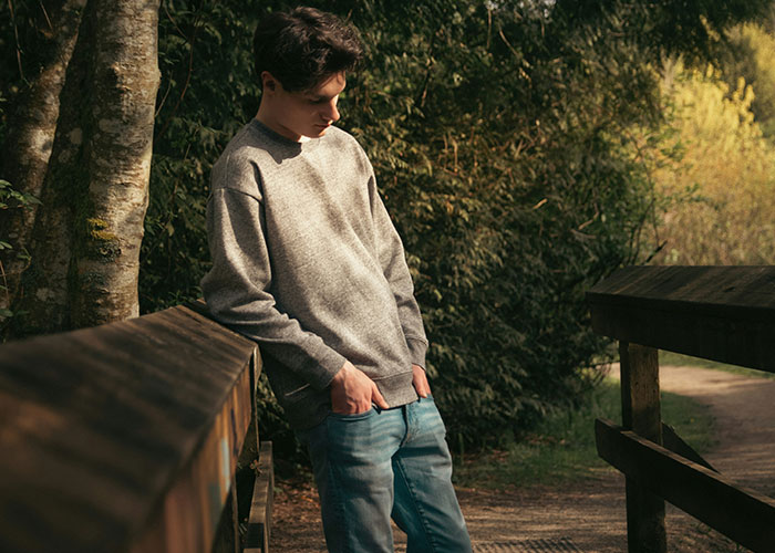 Teenager standing outdoors in casual clothes, looking down thoughtfully near a wooden fence surrounded by trees.