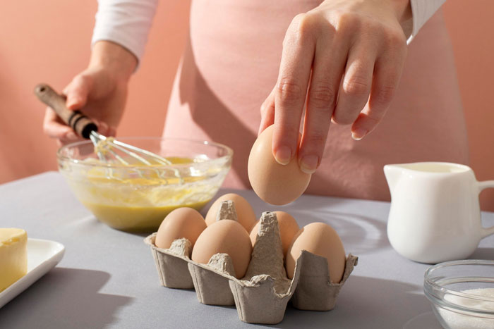 Hand holding an egg over carton with eggs beside a bowl of batter, illustrating eggs are dairy confusion in baking.