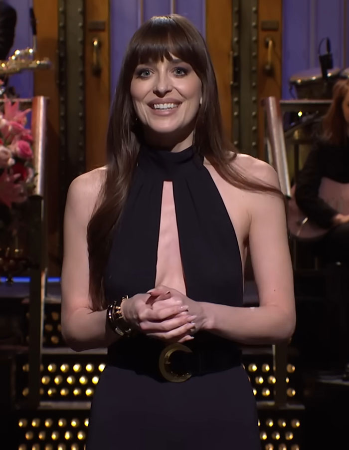 Dakota Johnson wearing a black sleeveless dress with a deep neckline, smiling while standing on stage at a live event. Dakota Johnson wearing a black sleeveless dress with a deep neckline, smiling while standing on stage at a live event.