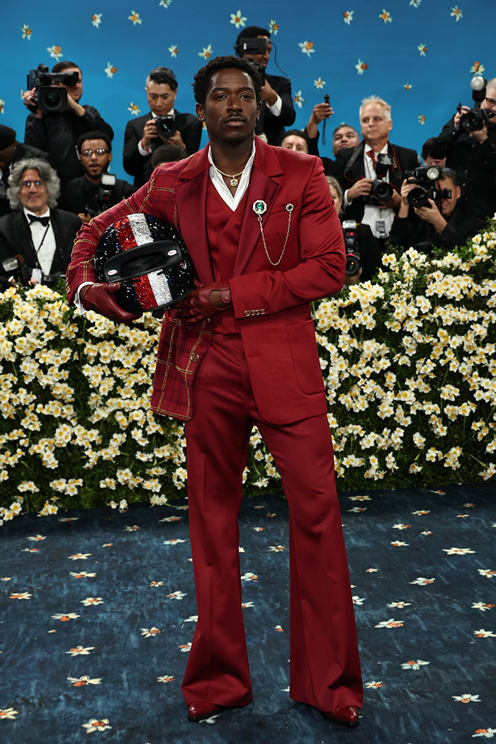 Damson Idris in a red suit holding a decorated F1 racing helmet, making a dramatic reveal at Met Gala 2025.