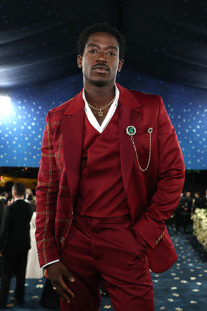 Damson Idris in a stylish red suit posing confidently at Met Gala 2025, hinting at dramatic reveal under F1 racing suit.