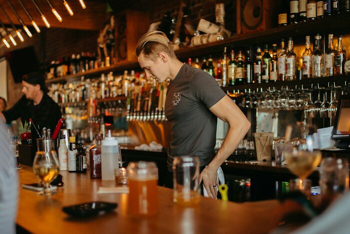 Bartender looking down thoughtfully behind a bar, illustrating moments from adult life feeling like being picked last in gym class.