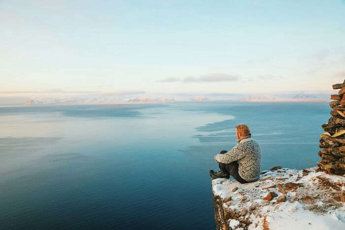 Person sitting on a snowy cliff overlooking a vast ocean, experiencing one of the must-see wonders of the world.