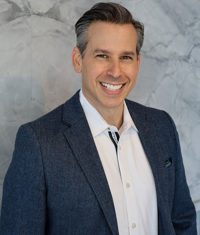 Smiling man in a blue blazer and white shirt posing against a marble background, related to Ozempic tongue side effect.