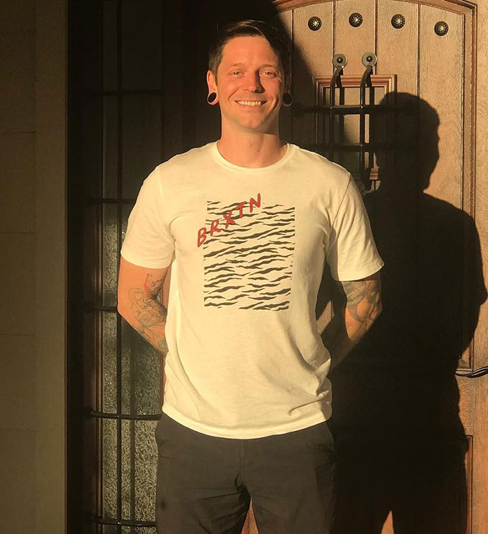 A famous drummer smiling, wearing a white graphic t-shirt, standing in front of a wooden door in warm sunlight.