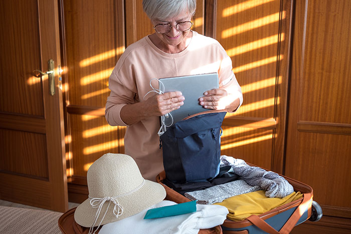 Older woman packing a suitcase for vacation, holding a tablet, showing mom booked a vacation and won't babysit kids.