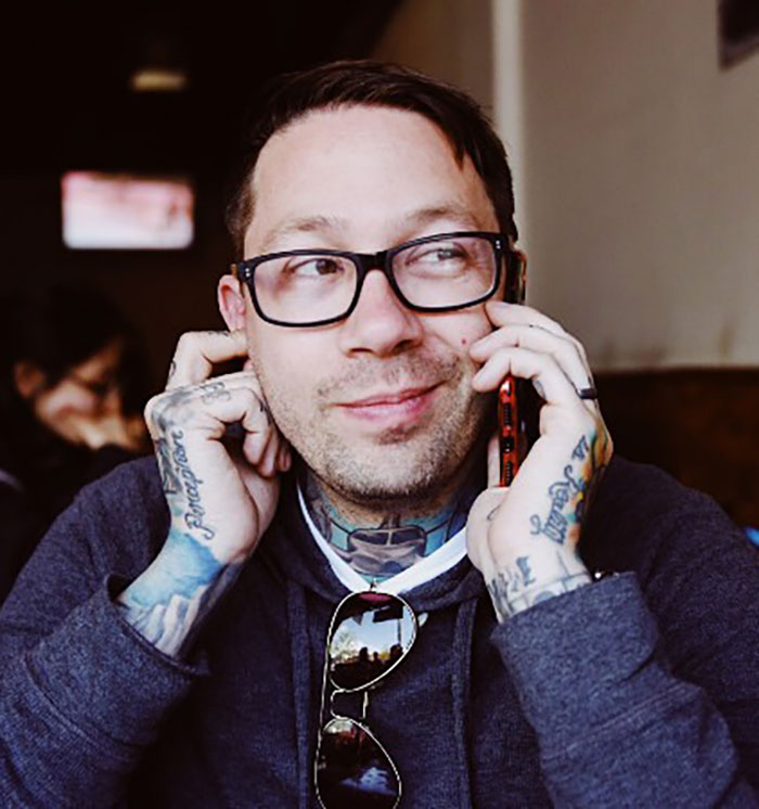 Famous drummer with tattoos and glasses sitting indoors, smiling while talking on phone after San Diego plane crash news.