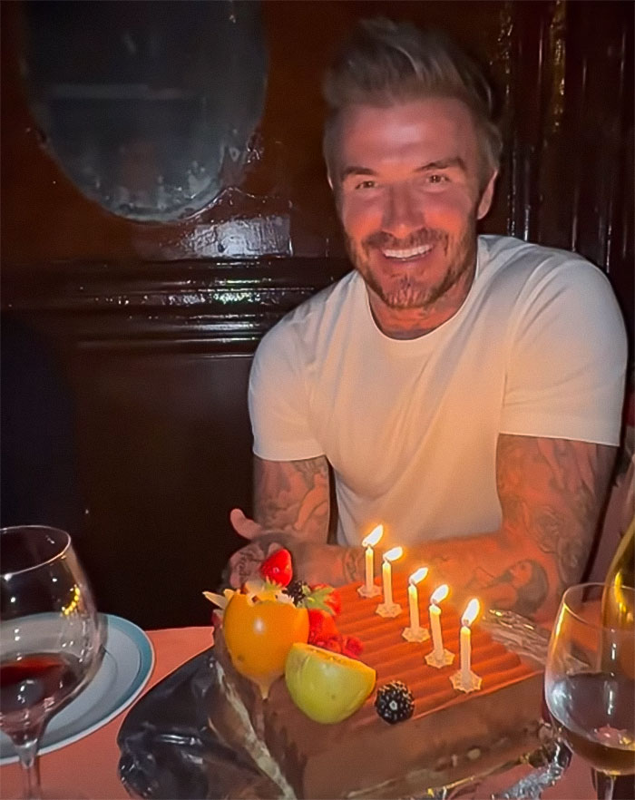 David Beckham celebrating with a birthday cake, smiling in a casual white shirt at his 50th birthday party. David Beckham celebrating with a birthday cake, smiling in a casual white shirt at his 50th birthday party.