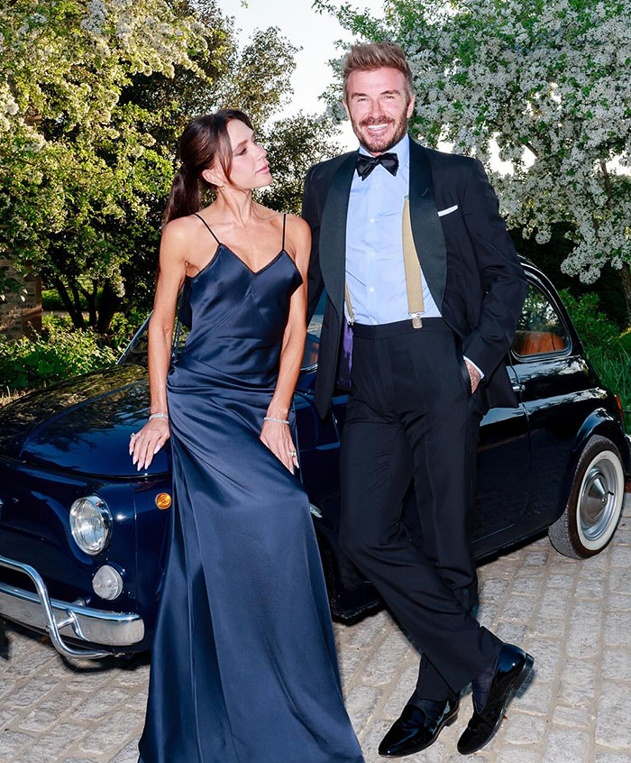 David Beckham and Victoria Beckham dressed formally posing by a vintage car at a 50th birthday party event.