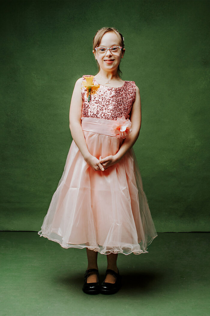 Young girl with Down syndrome in a pink dress smiling confidently against a green background, promoting inclusion.