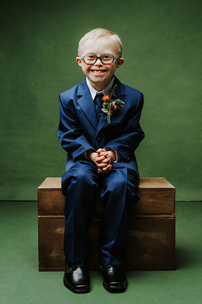 Young boy with Down syndrome in a blue suit and glasses smiling while sitting on a wooden box against a green background