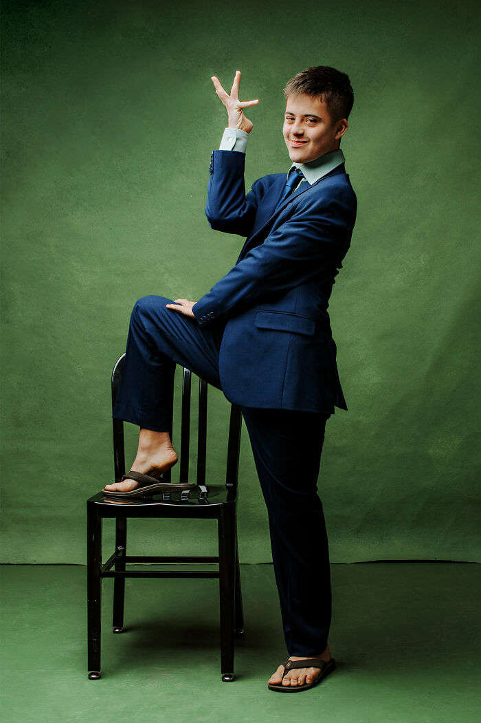 Young man with Down syndrome confidently posing in a blue suit and sandals against a green background, promoting inclusion.
