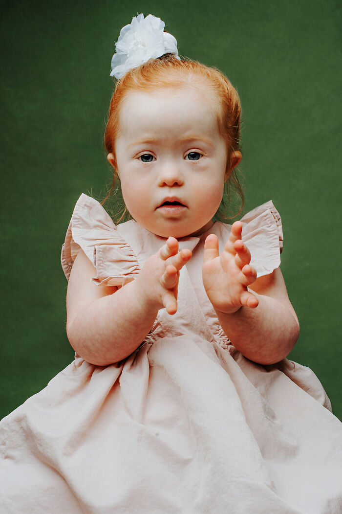 Young girl with Down syndrome wearing a light pink dress and white hair bow against a green background, celebrating inclusion.