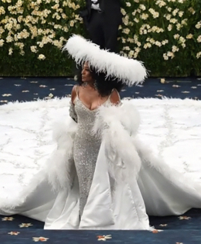 Zendaya in a sparkling gown and large feathered hat reacting on the Met Gala red carpet amid a floral backdrop.