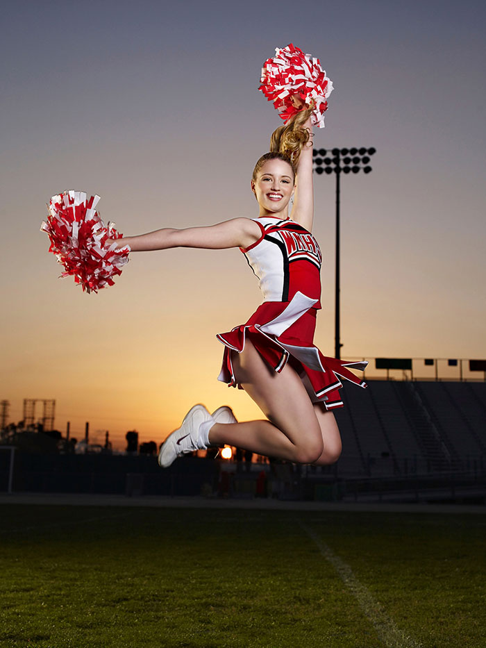Cheerleader jumping with red and white pom-poms at sunset on a sports field, showing energy and athleticism.
