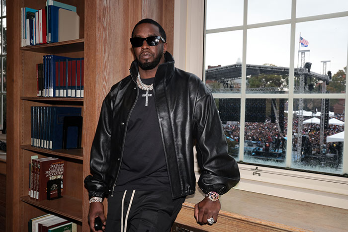 Diddy wearing a black leather jacket and sunglasses, posing indoors with a large crowd visible through the window behind him. Diddy wearing a black leather jacket and sunglasses, posing indoors with a large crowd visible through the window behind him.