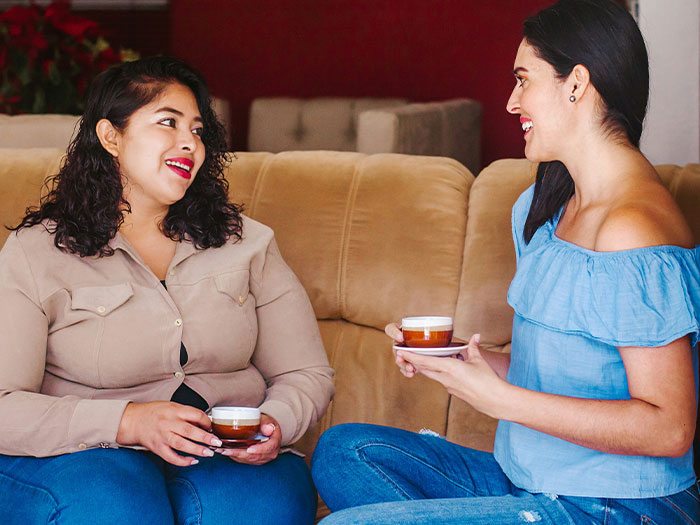 Two women having tea and smiling while discussing a distant cousin wanting funding for her wedding because of single status.