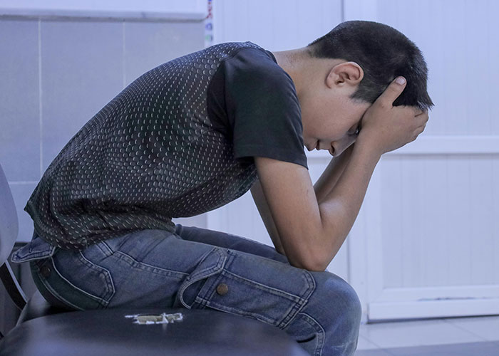 Teen boy sitting with head in hands, appearing distressed, illustrating dark family secrets and emotional struggle.