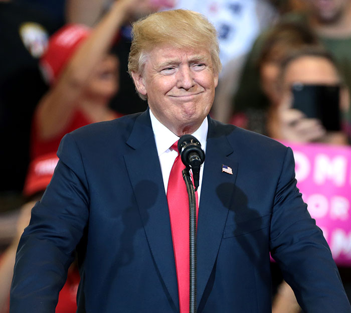 Former President Donald Trump speaking at a rally, in focus amid blurred enthusiastic crowd background.