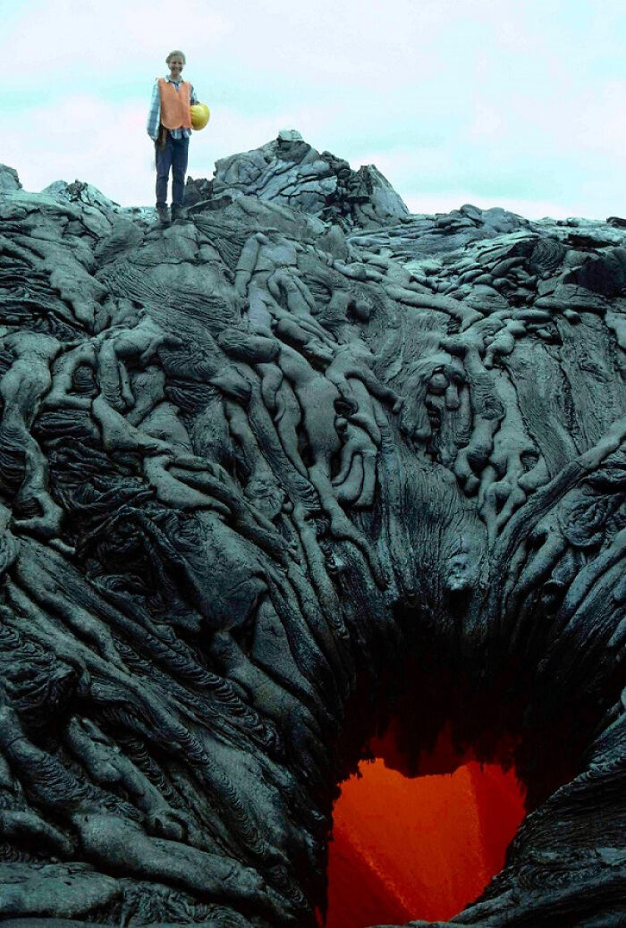 Person standing near a glowing lava hole surrounded by dark cooled lava rocks, capturing rare natural phenomena.