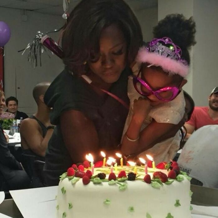 Woman and child wearing a tiara and oversized glasses blowing out candles on a birthday cake in a celebrity Instagram post.