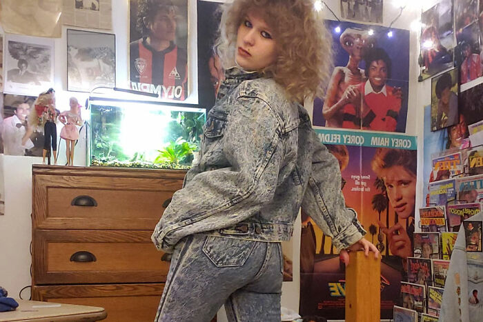 Person in acid-wash denim outfit posing in a retro room filled with vintage posters and collectibles, reflecting nostalgic fads.