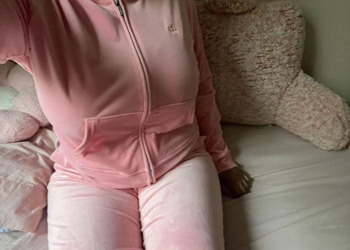 Person wearing a pink velour tracksuit sitting on a bed with a large teddy bear, reflecting on dumbest fad participated in.