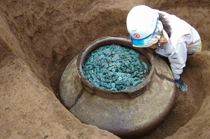 Archaeologist excavating a large ancient jar filled with numerous rare historical artifacts and coins in a dirt pit.