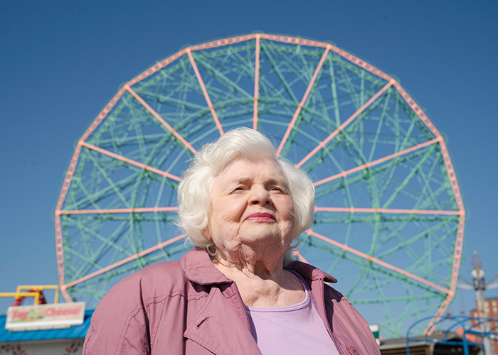 Elderly woman standing confidently with a Ferris wheel in the background on a clear day.