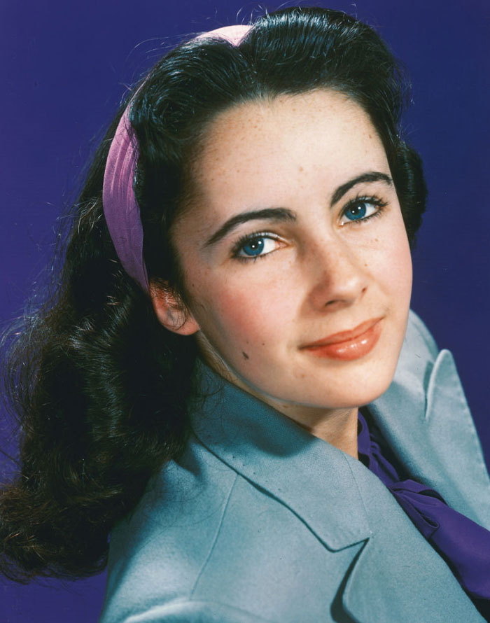 Young Elizabeth Taylor with striking blue eyes, wearing a purple headband and gray jacket against a dark blue background.