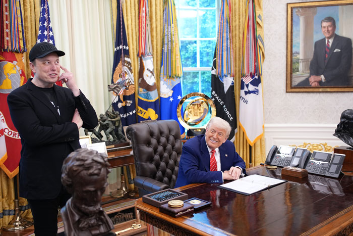 Elon Musk standing with a black eye and head ticks alongside a man seated at the Oval Office desk.