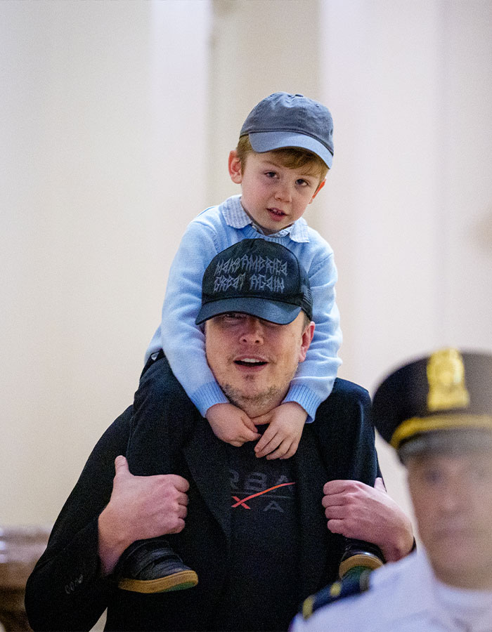 Elon Musk carrying a child on his shoulders, wearing a black cap, raising concerns after glitching and black eye appearance.