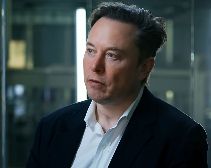 Elon Musk in a formal suit, speaking in an indoor corporate setting, related to Tesla CEO search news.