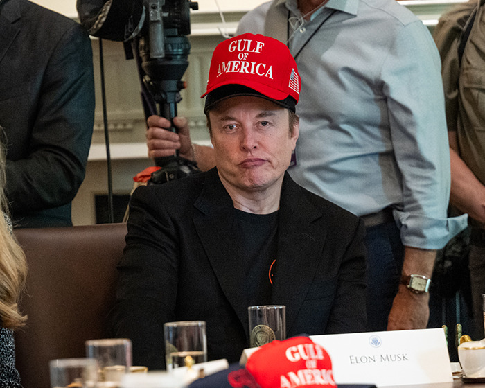 Elon Musk wearing a red Gulf of America hat, sitting at a meeting table with a serious expression. Elon Musk wearing a red Gulf of America hat, sitting at a meeting table with a serious expression.