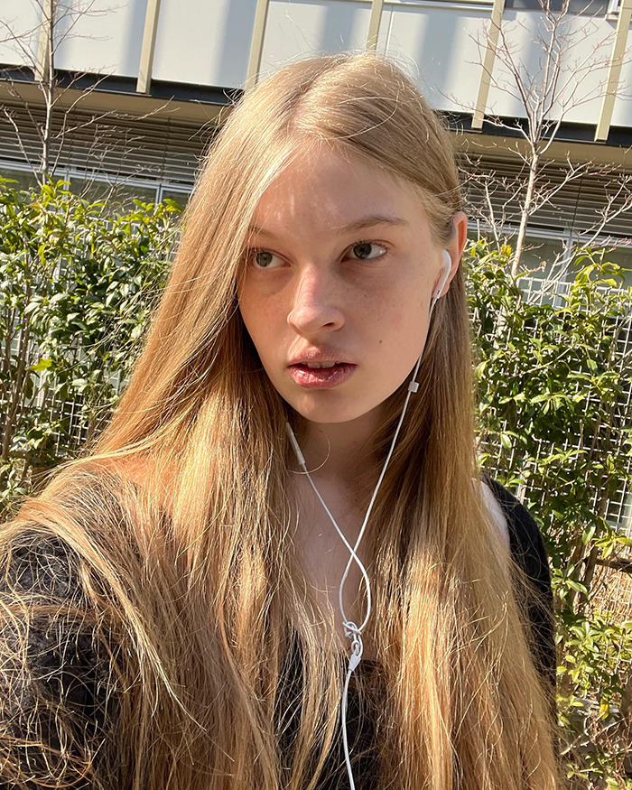 Elon Musk's trans daughter Vivian wearing earbuds outdoors with long blonde hair in a natural light setting.