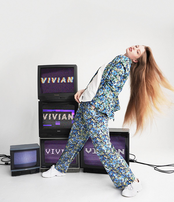 Vivian, Elon Musk's trans daughter, posing in a floral suit surrounded by vintage TVs displaying her name.