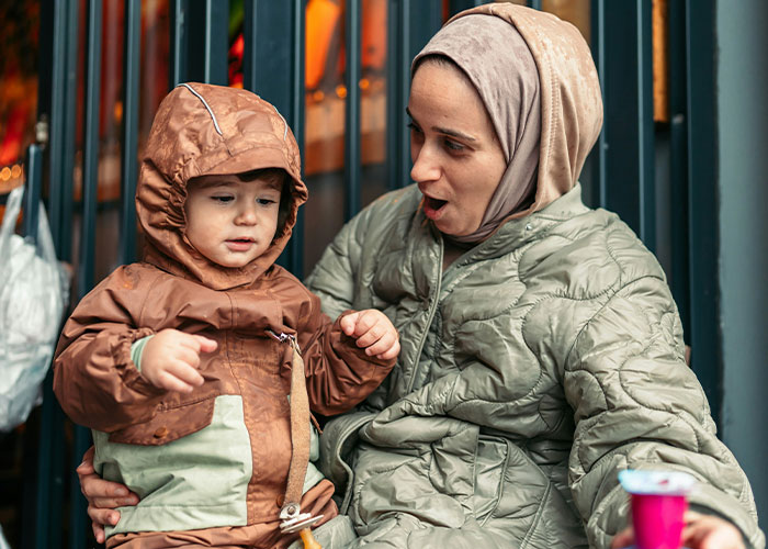 Mother wearing a headscarf looking shocked at her toddler in a brown raincoat in public, capturing parents’ kids’ worst moments.