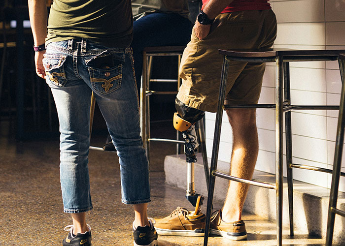Two people standing near bar stools, one wearing jeans and the other with a prosthetic leg, in a casual public setting.