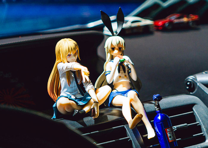 Two anime figurines, one with blonde hair and one with bunny ears, sitting on a car dashboard illustrating kids acting in public.