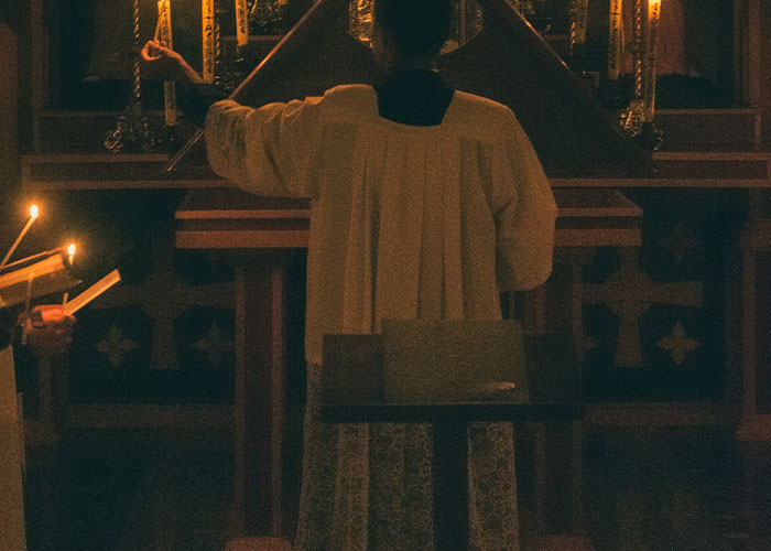 Person in religious robe raising hand in dimly lit church interior, illustrating parents sharing worst kids' public behavior.